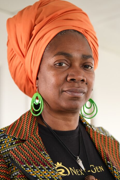 Photo of Torkwase Holmes by Dreph, in orange headscarf, green earrings and colourful jacket. 