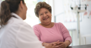 A woman speaking to a doctor. She is smiling and wearing a pink jumper.