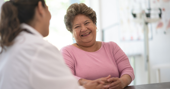 A woman speaking to a doctor. She is smiling and wearing a pink jumper.