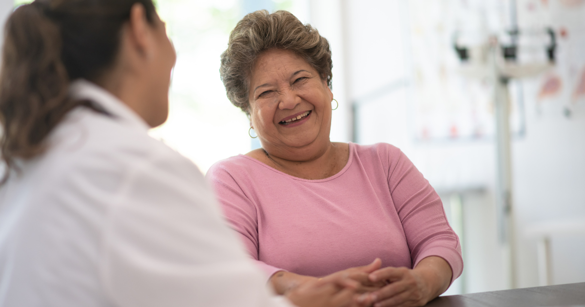 A woman speaking to a doctor. She is smiling and wearing a pink jumper.