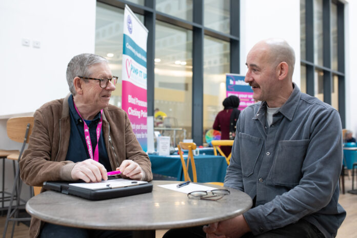 Two men sitting at a table completing a questionnaire. A Healthwatch banned is in the background.